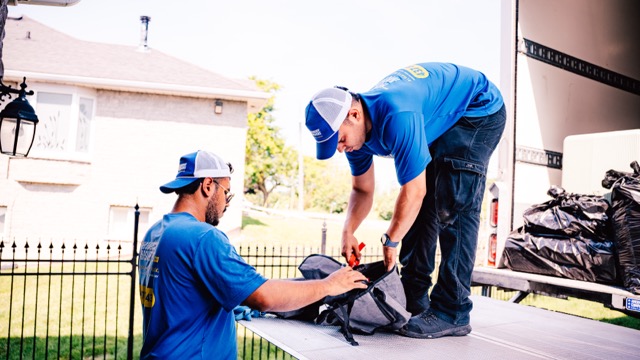 Two Homeboy Movers crew members loading a grey bag onto the truck ramp outside a house