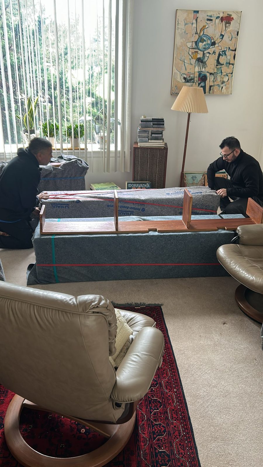 Two Homeboy Movers crew members wrapping and padding a sectional sofa in a living room